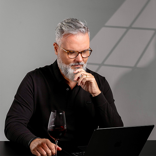 Foto de Alexandre de Salles em um ambiente profissional e sofisticado, analisando seu laptop enquanto aprecia uma taça de vinho tinto.